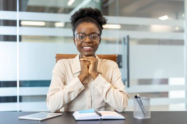Genç Afrikalı Amerikalı iş kadını masasında oturmuş, masadaki not defteri ve telefonuyla modern ofiste güvenle gülümsüyor. Cam duvarları ve rahat atmosferi olan profesyonel bir ortam..