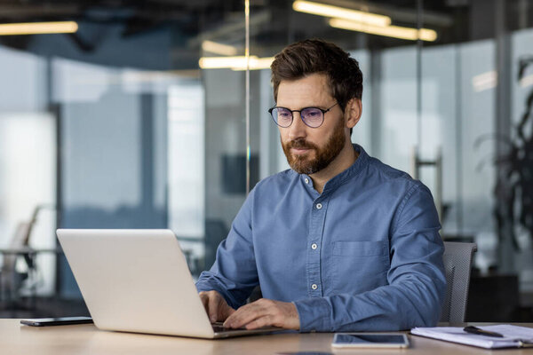 A young man in a shirt and glasses is working concentratedly in a modern office at a laptop, typing on a keyboard.
