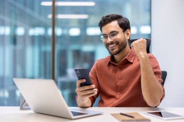 Heyecanlı iş adamı akıllı telefon kullanarak iş yerindeki başarısını kutluyor. Ofis masasında dizüstü bilgisayar, telefon ve defterle oturan genç profesyonel, neşeli ifade ve mutluluğu gösteriyor..