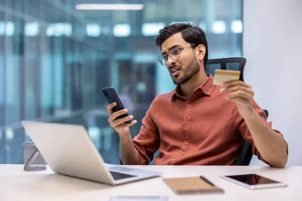 stock image Businessman in modern office holding credit card, looking concerned about phone fraud. Laptop, notebook, and tablet on desk. Concept of online fraud, cyber security, and technology issues