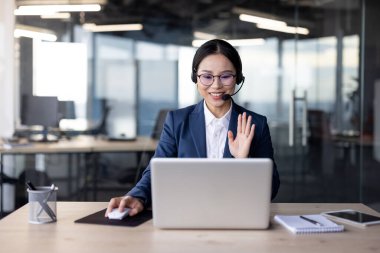 Modern ofis ortamında Asyalı bir iş kadını, kulaklık takıyor, bilgisayarında bir video görüşmesi yapıyor. Gülümsüyor ve ekranda el sallıyor. Profesyonel ve cana yakın görünüyor..