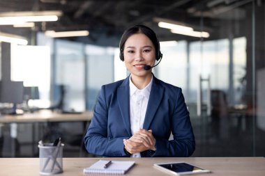 Modern bir ofis ortamında video aracılığıyla iletişim kuran resmi giyinmiş profesyonel bir iş kadını. Kameraya bakıyor ve kulaklık takıyor. Profesyonellik taslıyor.