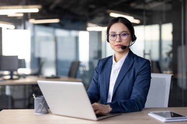 Kulaklık takan Asyalı iş kadını modern ofis ortamında dizüstü bilgisayar üzerinde çalışıyor. Profesyonel ortam verimliliği ve kararlılığı yansıtır.