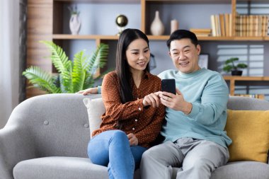 Mutlu Asyalı çift kanepede oturmuş akıllı telefon kullanıyor, internete bakıyorlardı. İfadeleri, konforlu ev ortamında teknolojiyle neşe ve bağlantı gösteriyor.