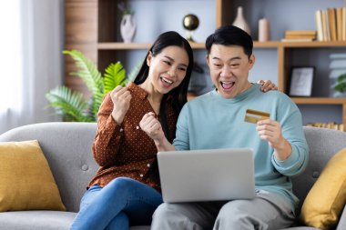 İnternetten alışveriş yapmak için dizüstü bilgisayar ve kredi kartı kullanan Asyalı mutlu bir çift. Gülümseyen ikili başarılı alışverişi kutluyor, birlikte vakit geçiriyorlar. E-ticaret ve teknoloji kavramı