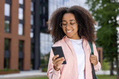 Güneşli bir günde açık havada akıllı telefon kullanan gülümseyen Afrikalı Amerikalı kadın. Kıvırcık saçlı, gözlüklü, telefon ekranına bakan kendine güvenen genç bir kadın. Gelişigüzel kentsel ortam