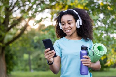 Parkta yoga minderi ve su şişesi tutarken gülümseyen Afro-Amerikalı kadın. Sağlıklı yaşam tarzını ve modern teknolojiyi vurgulayan açık hava fitness ve sağlık konsepti