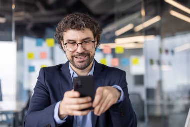 Modern ofiste gözlük ve takım elbise takan kendine güvenen bir işadamı telefon kullanır. Çağdaş iş ortamında çalışan akıllı telefonlu mutlu profesyoneller.