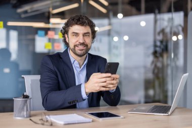 Modern ofiste akıllı telefon kullanan kendine güvenen iş adamı kameraya gülümsüyor. Profesyonel kıyafet, dizüstü bilgisayar, masa başı not defteri. Üretkenlik ve işbirliği için ideal, parlak, profesyonel çevre.