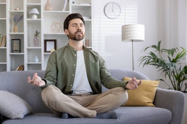 Oturma odasındaki kanepede meditasyon yapan, farkındalık ve rahatlama egzersizi yapan bir adam. Evde sakin ve iç huzuru koruyan kişi, meditasyon pozisyonunda gözleri kapalı.