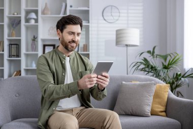 Evdeki kanepede otururken tablet kullanan gülümseyen adam. Modern teknolojiden hoşlanan mutlu bireyler, boş zamanlarında ya da uzak işlerde çalışırlar. İçerisi aydınlık ve rahat. Android ve iOS uyumlu.