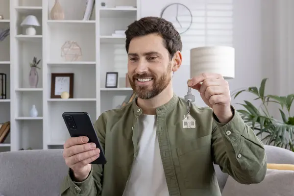 stock image Smiling man holding new house keys while looking at phone. Depicts excitement, real estate success, new home ownership, and modern lifestyle. Concept of achievement and digital communication.