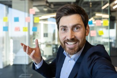 Kendine güvenen işadamı, modern ofisteki telefon görüşmesinde gülümsüyor ve jest yapıyor. Takım elbiseli profesyonel iş arkadaşları veya müşterilerle sanal toplantı yapıyor..