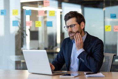 Masada oturan profesyonel bir iş adamı dizüstü bilgisayarla çalışırken diş ağrısı çekiyor. Ofis ayarları, cam duvarda yapışkan notlar ve masanın üstünde telefon..
