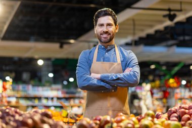 Önlük takan kendine güvenen erkek market çalışanı üretim koridorunda kolları çapraz duruyor. Taze meyve ve sebzeler ön planda. Parlak mağaza ışıkları sıcak, davetkar atmosfer yaratır