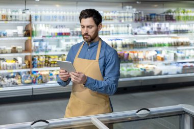 Bej önlüklü bir market çalışanı süpermarkette envanter yönetimi için tablet kullanıyor ve raflarda çeşitli ürünler bulunuyor. İşçi iş operasyonları için teknolojiye odaklandı