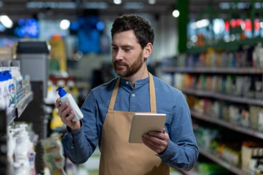 Süpermarket çalışanı envanteri kontrol ederken elinde ilaç şişesi ve tablet tutuyor. Perakende mağazası koridorunda çalışan önlüklü bir adam hisse senedi kontrolü ve yönetimi için malları inceliyor..