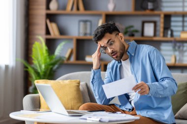 Adam evdeki mali belgeleri incelerken endişeli görünüyor. Dizüstü bilgisayarda çalışıyor ve evrakları tutuyor. Finansal stres, bütçe ve ev ofisi kurulumu kavramı.