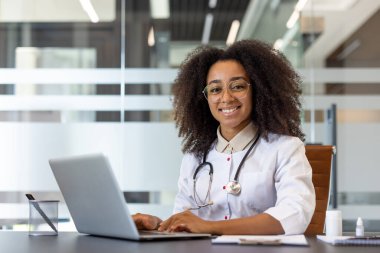 Gülümseyen bir doktor elinde dizüstü bilgisayarla bir masada oturmuş modern ve iyi aydınlatılmış bir ofiste online danışmanlık öneriyor. Atmosfer profesyonelce ve doktor hastalara yardım etmeye hazır..