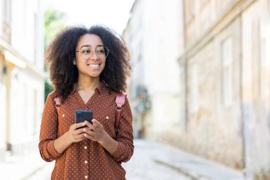Kıvırcık saçlı ve gözlüklü bir kadın turist güneşli bir caddede geziniyor, tatilinin tadını çıkarıyor. Elinde bir akıllı telefon, kahverengi bir gömlek ve pembe bir sırt çantası var..