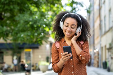 Gülümseyen kadın şehir caddesinde akıllı telefon kullanırken kulaklıkla müzik dinlemekten zevk alıyor. Rahatlamış ve mutlu. Cihazını açık havada kullanıyor..
