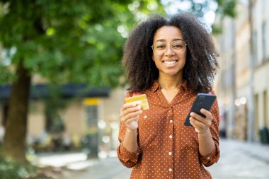 Açık havada kredi kartıyla akıllı telefonu online alışveriş için kullanan gülümseyen bir kadın. Şehir ortamında kendinden emin, mutlu ifade ve modern teknoloji kullanımı yoluyla gösterilen dijital ödeme kavramı.