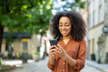 Şehrin ortasında dikilirken akıllı telefon kullanan gülümseyen bir kadın. Arka planda yeşil ağaçlarla aydınlık güneşli bir gün. Kentsel yaşam tarzı ve iletişim konsepti.