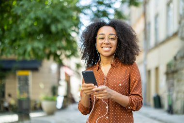 Açık havada kulaklıkla müzik dinlerken akıllı telefon kullanan kıvırcık saçlı, gülümseyen bir kadın. Mutlu yüz ifadesi, açık hava aktivitelerinden ve gözlüklü günlük kıyafetlerden keyif alıyor..