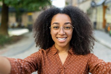 Kıvırcık saçlı ve gözlüklü mutlu kadın güneşli bir günde selfie çekiyor. Açık havada duruyor, kahverengi noktalı bir tişört giyiyor ve kameraya gülümsüyor. Doğal arka plan bulanık sokak manzarası.