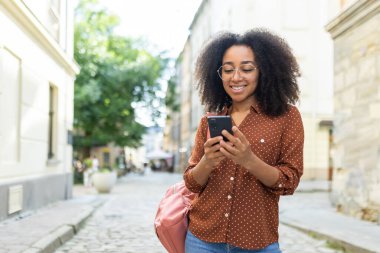 Güneşli bir sokakta yürürken akıllı telefonunu kullanan kıvırcık saçlı ve gözlüklü neşeli bir kadın. Gününün tadını çıkarıyor, bağlanıyor ve telefonuyla meşgul..