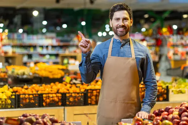 Önlüklü bir market çalışanı sebze reyonunda dikilip işaret ediyor. Sergilenen parlak meyve ve sebzeler renkli bir arkaplan oluşturuyor. Müşteri hizmetleri, perakende, gıda ve pazarın resmi.