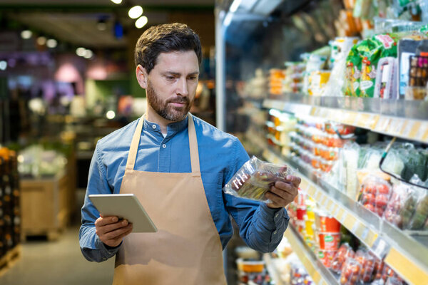 Grocery store worker holding tablet checking product on shelf in supermarket. Supermarket employee conducting inventory, ensuring product quality. Retail management and store operations concept.