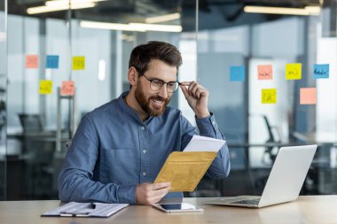 Gülümseyen işadamı büro masasında iyi haberlerle mektup okuyor. Profesyonel gözlüklü, zarflı, heyecanlı. Arka planda teknoloji ve ofis notları.
