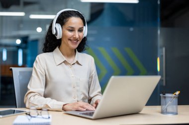 Kendine güvenen iş kadını kulaklık takıyor, dizüstü bilgisayarla gülümsüyor. Modern ofis ortamında profosyonelliği, teknolojiyi ve iletişimi destekleyen üretken bir işe giriştim.