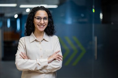 Modern ofis ortamında gülümseyen gözlüklü kendine güvenen genç bir kadın. Mesleki kıyafet, olumlu davranış, işyerindeki başarı ve şevki yansıtıyor. Mesleki motivasyon için ilham.