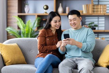 Kanepede oturan Asyalı çift telefona bakarken keyifli anlar yaşıyor. Kadın ve erkek, modern oturma odasında gülümsüyor ve kaynaşıyorlar..