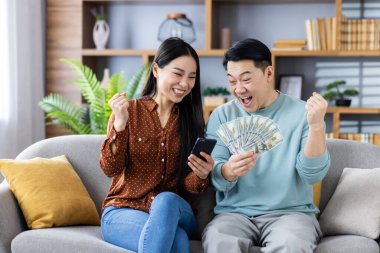Neşeli Asyalı çift finansal başarıyı nakit ve telefonla kutluyor, evdeki kanepede oturuyor, heyecanlı hissediyorlar. Finansal başarı, mutluluk ve birliktelik kavramı.