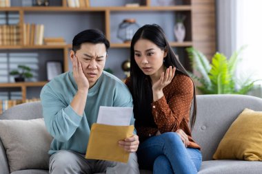 Kanepede oturan Asyalı çift faturaları kontrol ederken stresli ve endişeli görünüyor. Erkek ve kadın mektupları ve mali belgeleri endişeyle inceliyor.