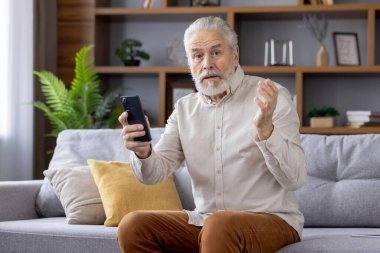 Hüsrana uğramış ve hayal kırıklığına uğramış üzgün bir adamın portresi. Kanepede oturmuş, elinde telefonla üzgün bir şekilde kameraya bakıyor ve ellerini açıyor..