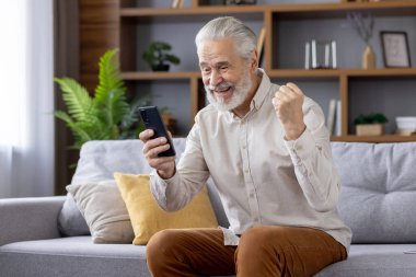 Mutlu kıdemli adam zaferin tadını çıkartıyor. Elleriyle zafer işareti yapıyor, koltukta oturuyor ve cep telefonu kullanıyor..