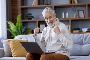 Yaşlı, kır saçlı adam evinde, kanepede oturuyor, kucağında dizüstü bilgisayar tutuyor ve video iletişiminin kamerasına bakıp mutlu bir şekilde bakıyor, zafer işareti yapıyor elleriyle..