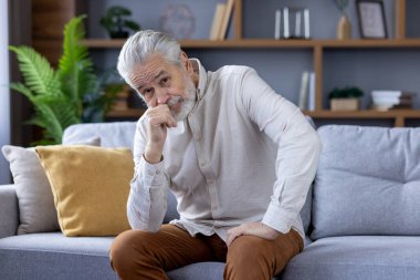 Ciddi ve düşünceli bir kıdemli adamın portresi kanepede oturuyor, başını eline yaslıyor ve üzgün bir şekilde kameraya bakıyor..