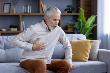 Yaşlı, kır saçlı adam evdeki kanepede oturuyor ve elini göğsüne dayayıp kalp krizi ve kalp krizi geçiriyor..