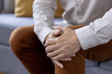 Vücut fotoğrafı, kahverengi pantolonla dizini tutan yaşlı bir adamın elleri, şiddetli bir acı ve burkulma hissi..