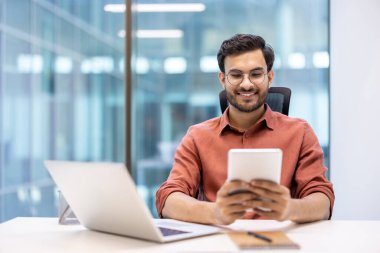 Modern ofiste tablet ve laptop kullanan gülümseyen bir programcı. Verimlilik ve konsantrasyon gösteren dijital cihazlarla uğraşan adam.