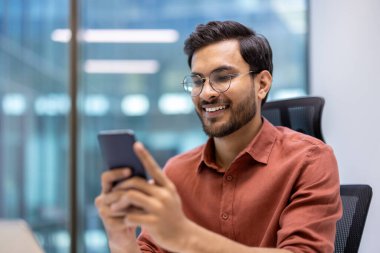 Ofis ortamında gözlüklü ve akıllı telefon kullanan neşeli bir adam. Telefonla konuşurken mutlu ve rahat bir ifade. Turuncu gömlekli profesyonel bir kıyafet.