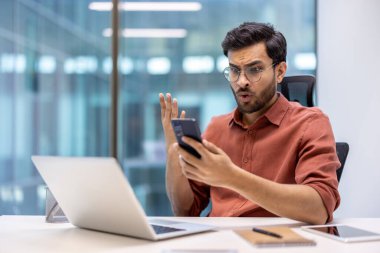 Modern ofiste oturan adam dizüstü bilgisayarla çalışırken telefon mesajına şaşırmış görünüyor. İfade şok ve güvensizlik gösteriyor. Dijital aygıtlar ve çalışma alanı olan ofis ortamı.