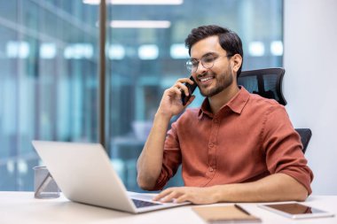 Gülümseyen işadamı dizüstü bilgisayarla çalışırken telefonda konuşuyor. Proje, çoklu görev ve bağlı kalma üzerine profesyonel çalışma ile ofis ortamı.