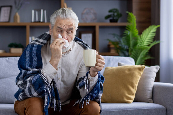 Пожилой человек плохо себя чувствует, сидит дома на диване, покрытом одеялом, держит чашку с напитком в руках и вытирает нос салфеткой из насморка..
