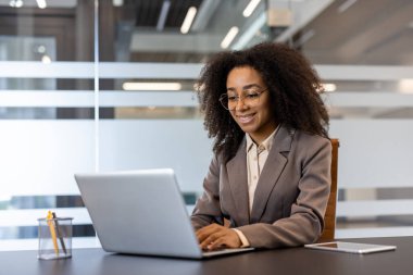 Genç Afrikalı Amerikalı iş kadını modern ofiste dizüstü bilgisayar kullanıyor. İş yerinde bilgisayarla çalışan mutlu ve kendinden emin bir kadın. Dizüstü bilgisayarda gülümseyen profesyonel ve başarılı bir çalışan.